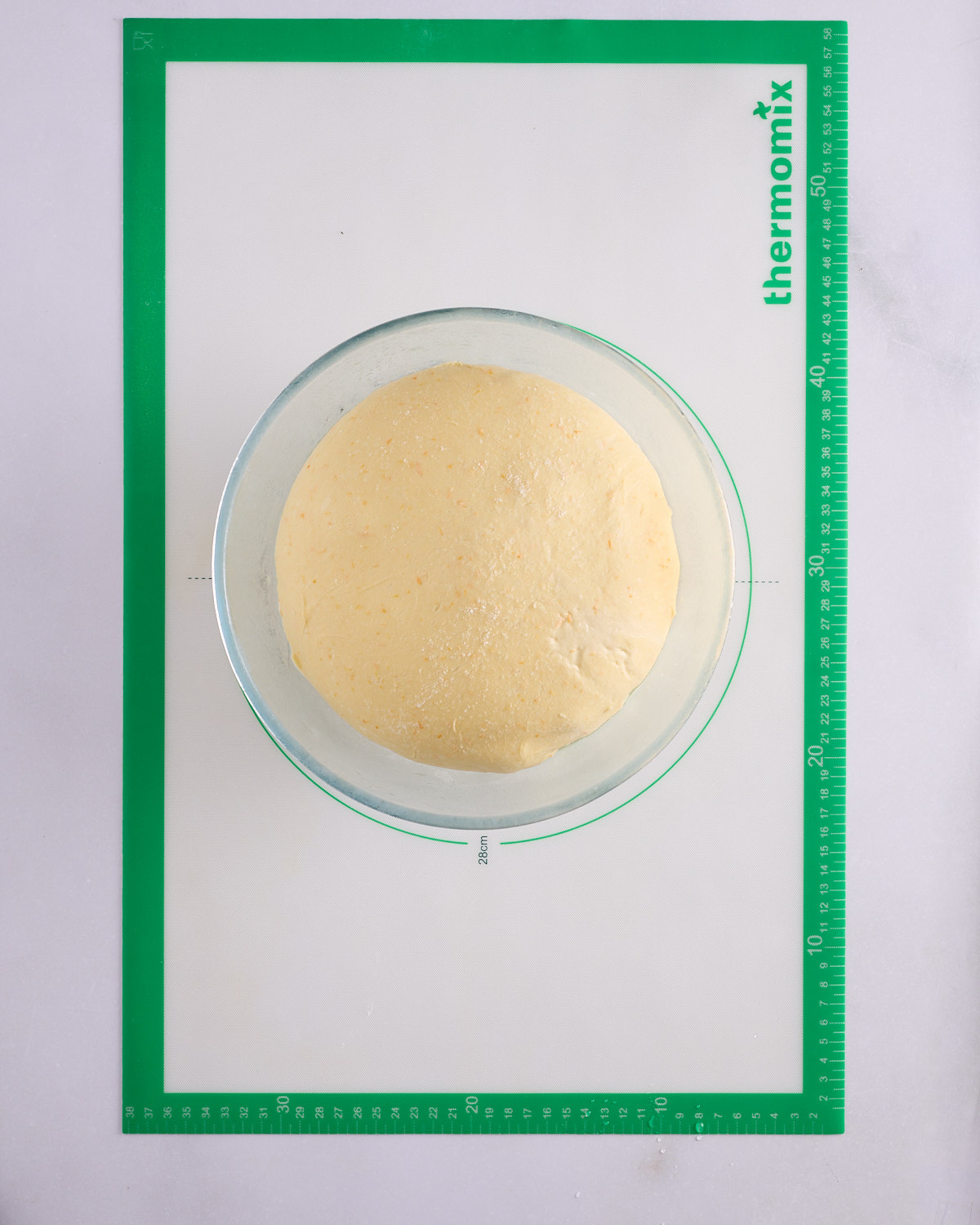 Risen foundation sweet dough in a glass bowl on a green Thermomix pastry mat, the dough measuring about 28cm across.
