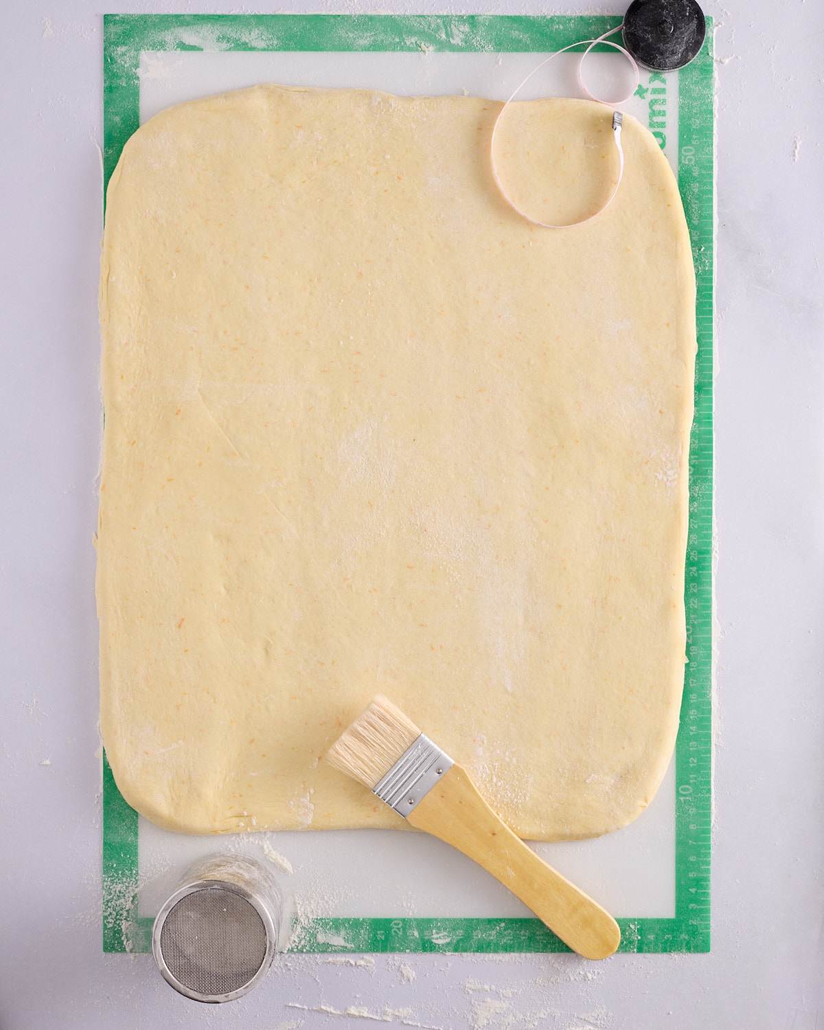The foundation sweet dough rolled into a large rectangle on a green silicone mat, flecked with orange zest, with a pastry brush, flour sifter and dough cutter arranged around it.