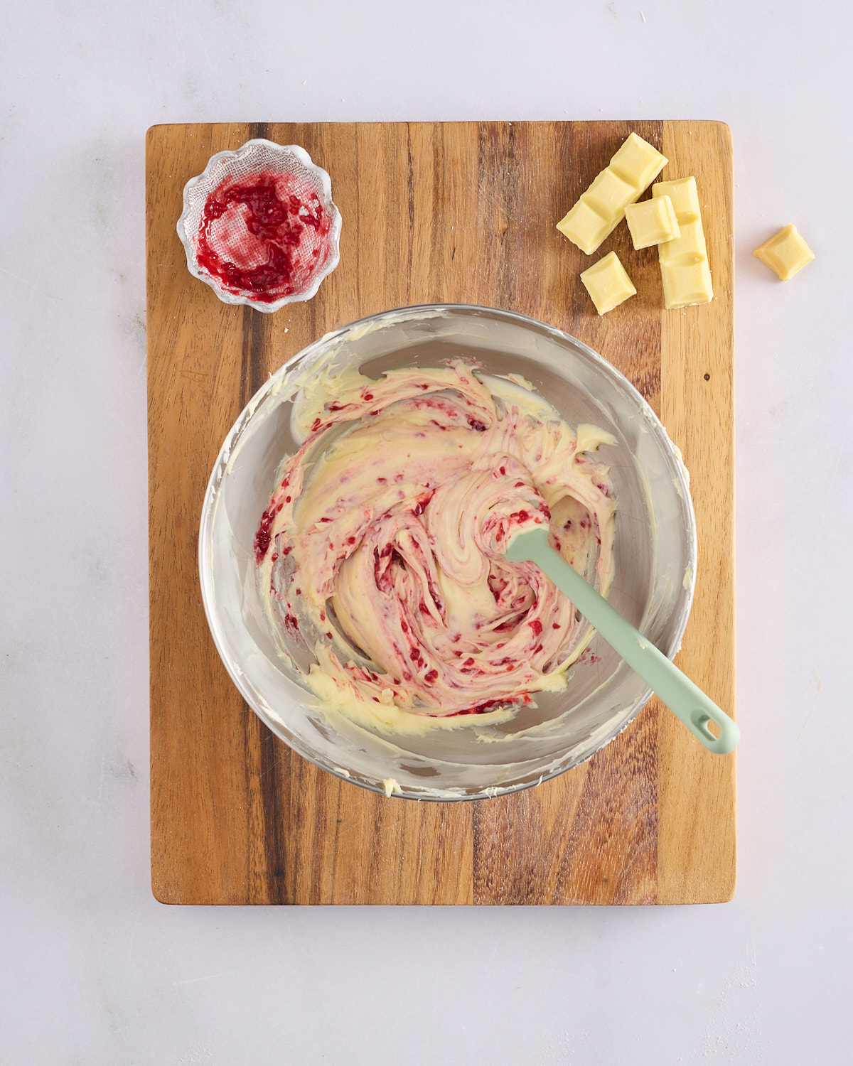 A stand mixer bowl of pale cream cheese frosting being marbled with raspberry jam by hand with a silicone spatula, on a wooden board, with a small dish of raspberry jam and chunks of white chocolate alongside.