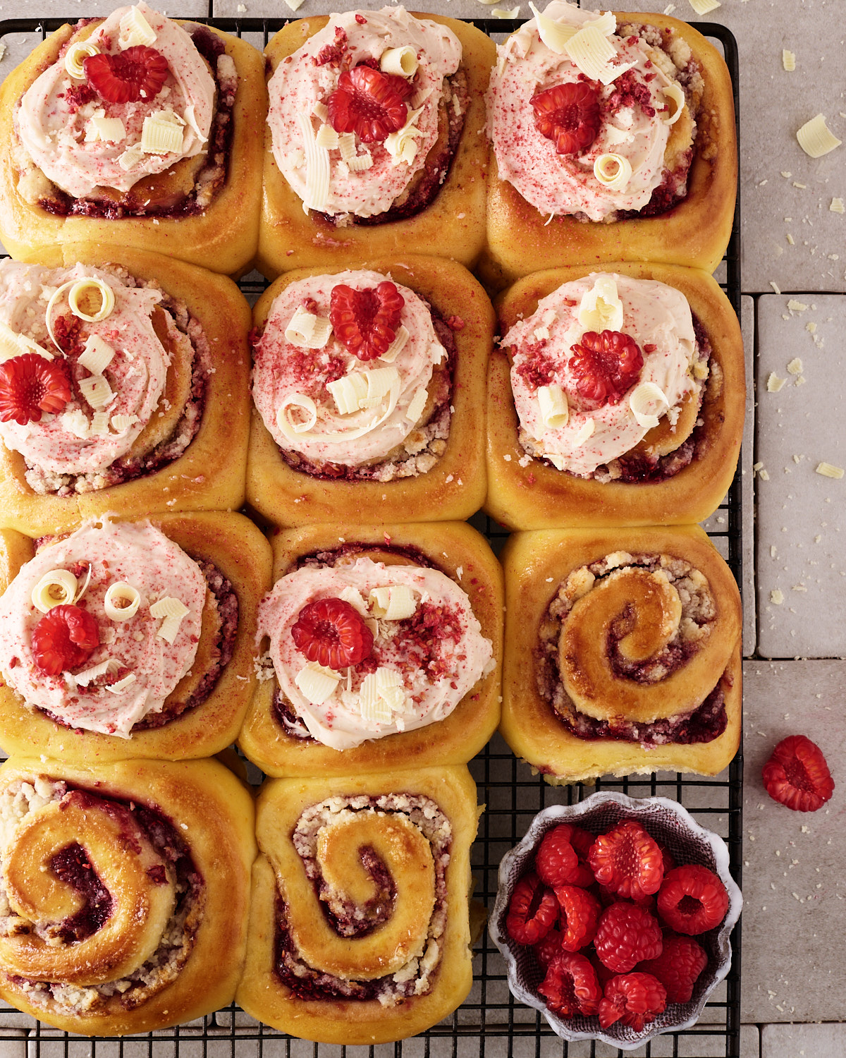 Twelve raspberry macadamia scrolls on a black wire cooling rack, eight topped with pink raspberry white chocolate cream cheese frosting, fresh raspberries, white chocolate curls and freeze-dried raspberry, and four left unfrosted to show the spiral of low sugar raspberry jam and macadamia marzipan, with a small dish of fresh raspberries alongside.
