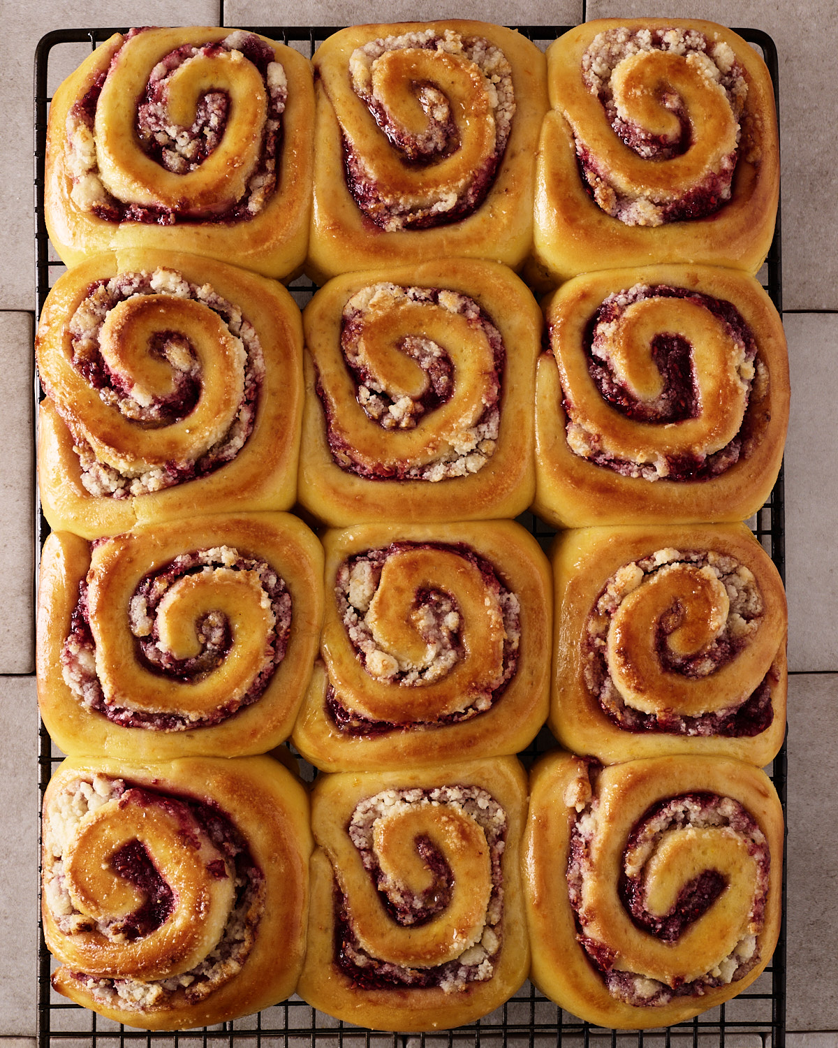 Twelve baked raspberry macadamia scrolls in a four-by-three grid on a black wire cooling rack, golden and glossy from the egg wash, each showing a spiral of raspberry jam and macadamia marzipan, on a tiled surface.