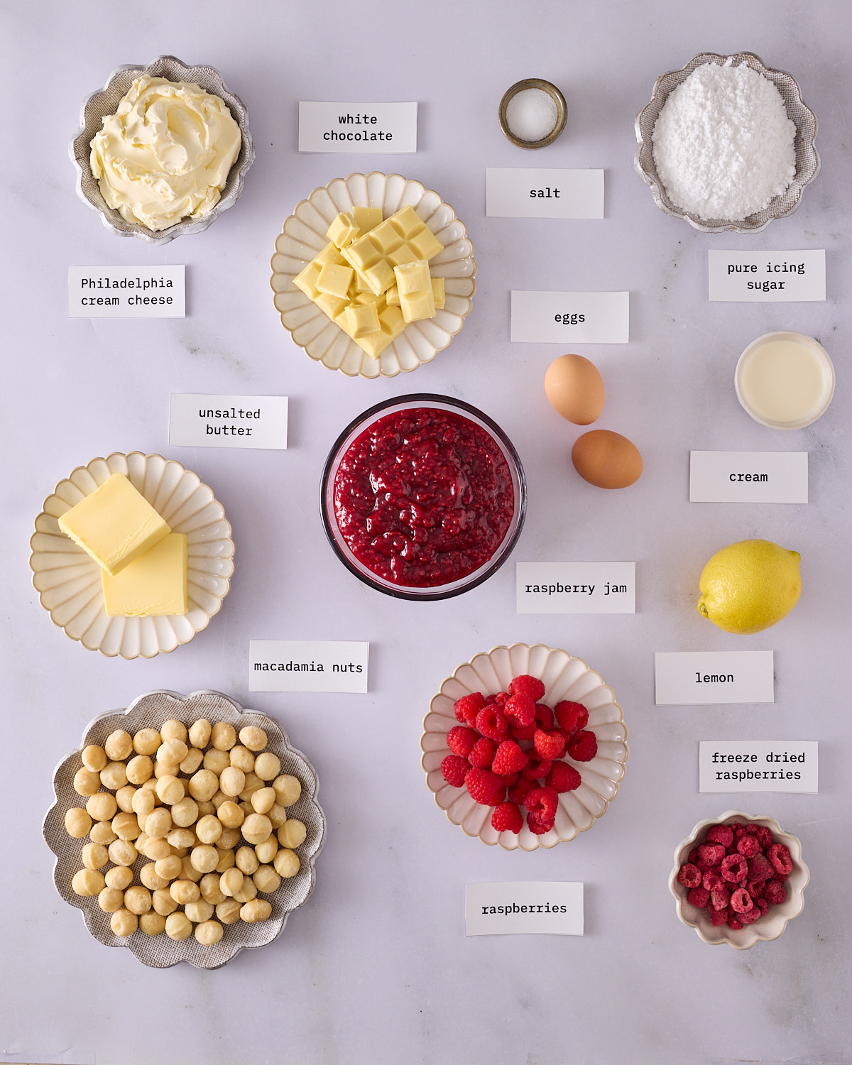 Labelled ingredients for the filling, frosting and décor for raspberry macadamia scrolls on a marble surface. Philadelphia cream cheese, white chocolate, raspberry jam, macadamia nuts, fresh raspberries, freeze dried raspberries, pure icing sugar, two eggs, cream, a lemon and salt.