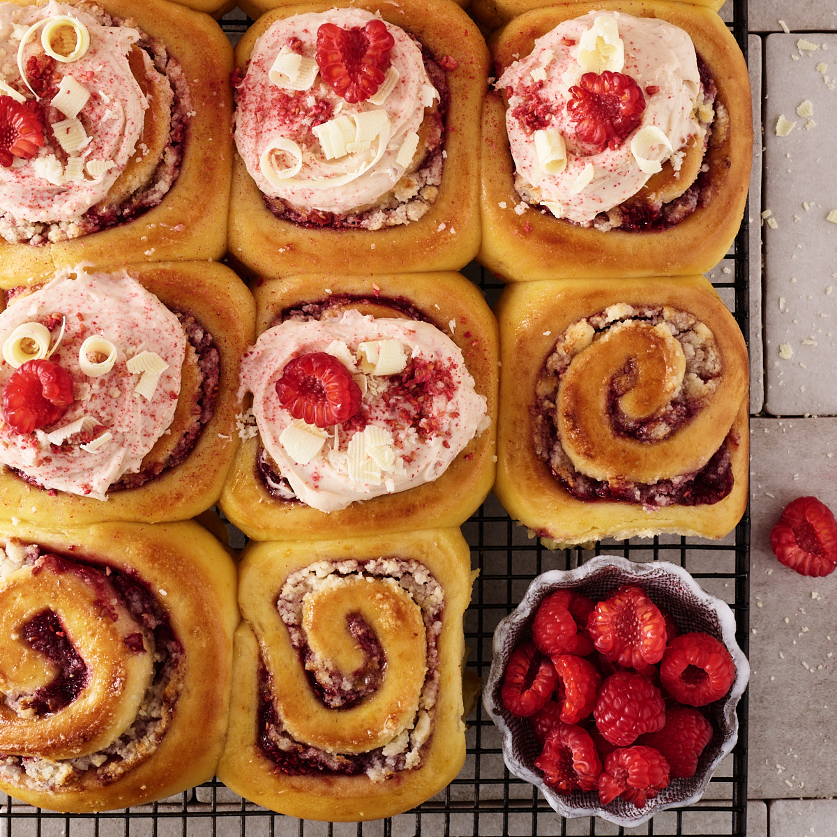 Twelve raspberry macadamia scrolls on a black wire cooling rack, eight topped with pink raspberry white chocolate cream cheese frosting, fresh raspberries, white chocolate curls and freeze-dried raspberry, and four left unfrosted to show the spiral of low sugar raspberry jam and macadamia marzipan, with a small dish of fresh raspberries alongside.