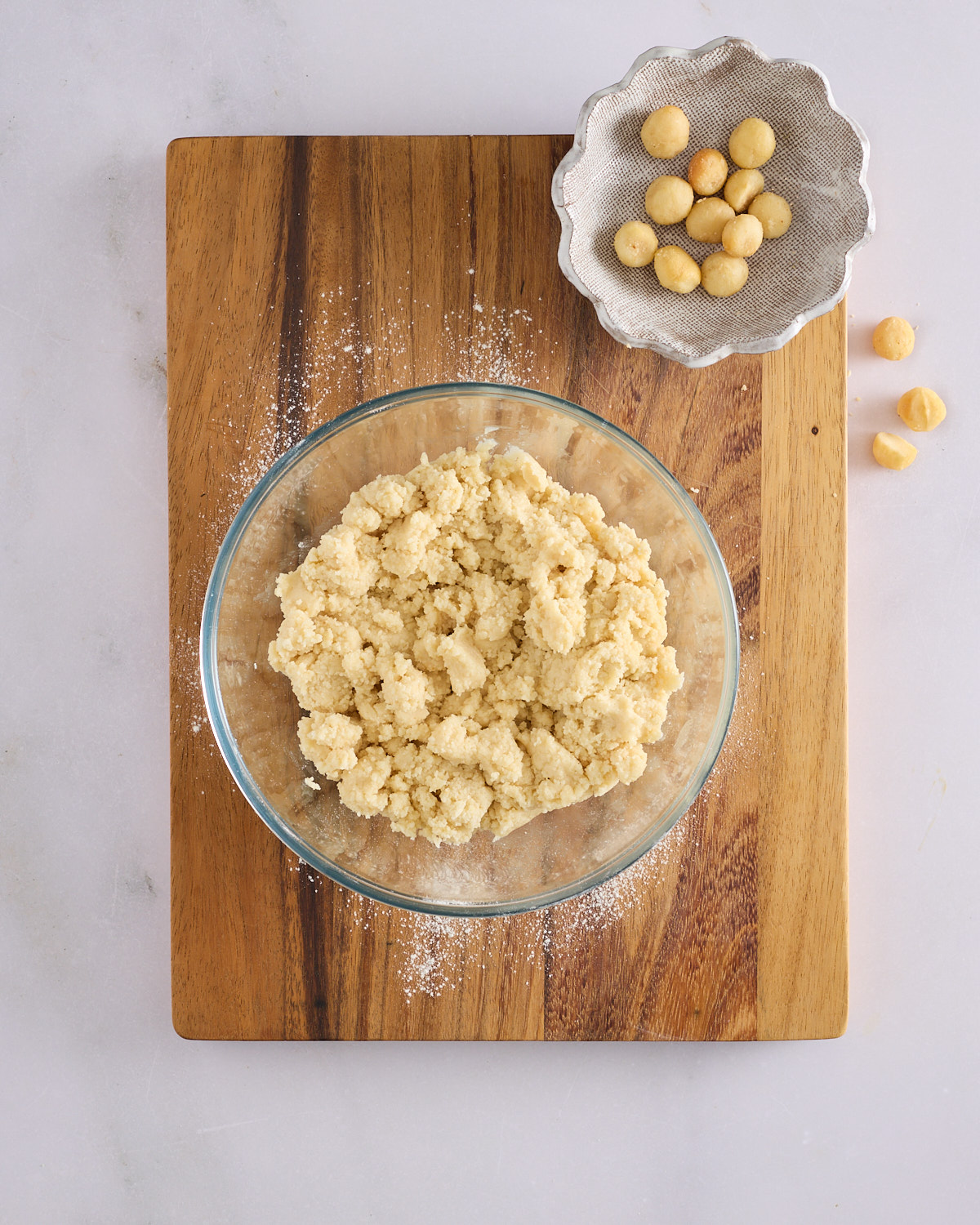 Crumbled macadamia marzipan in a glass bowl on a wooden board, with a dish of roasted macadamia nuts alongside.