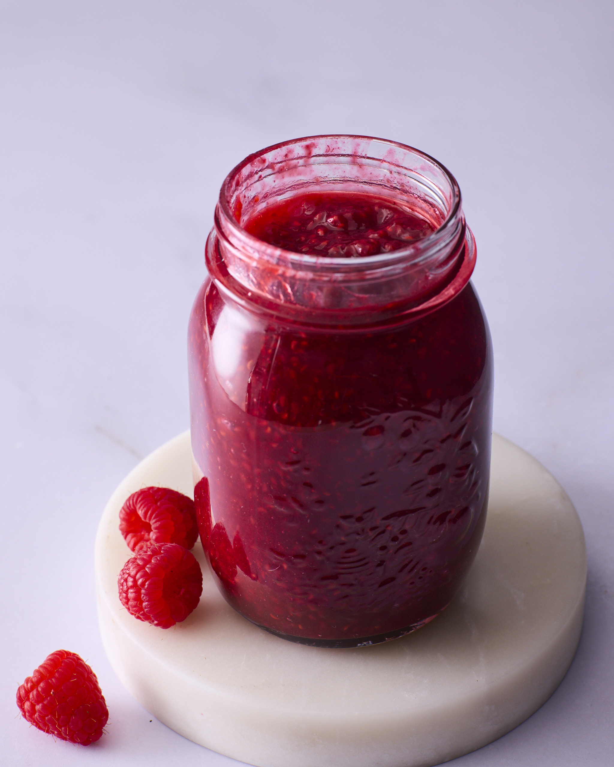 A jar of homemade raspberry jam on a round marble board, with three fresh raspberries beside it against a pale background.