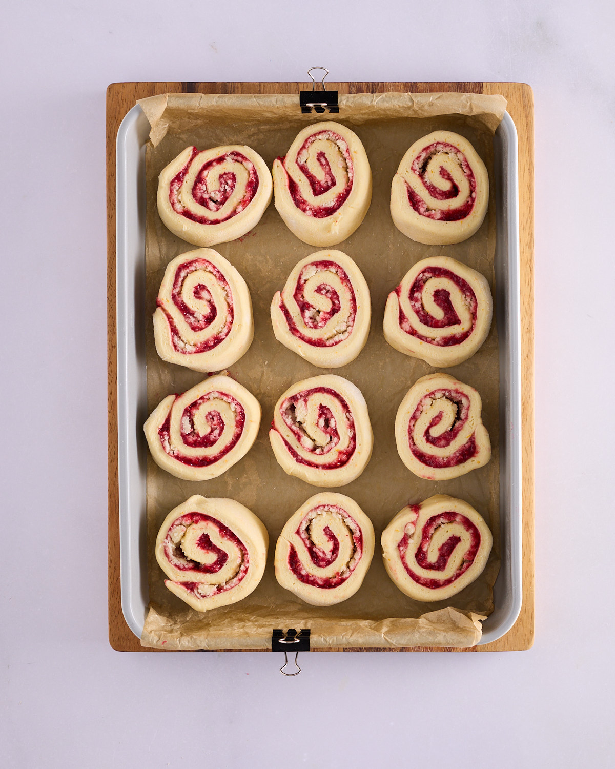 Twelve raspberry macadamia scrolls arranged three across and four down in a parchment-lined baking pan before proving, each showing a spiral of raspberry jam and macadamia marzipan, on a wooden board.